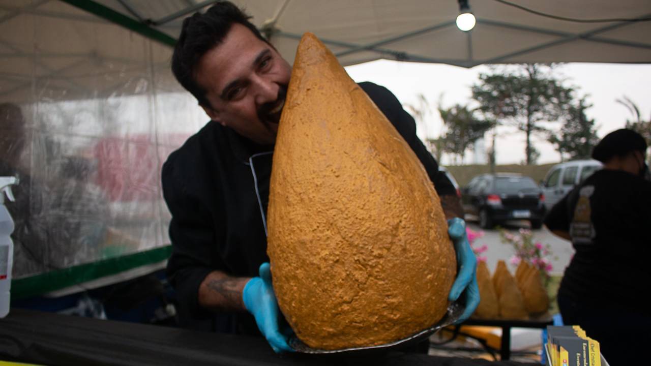 Homem posa em barraca usando luvas azuis e segurando coxinha enorme, do tamanho de seu torço, e fingindo dar uma mordida.