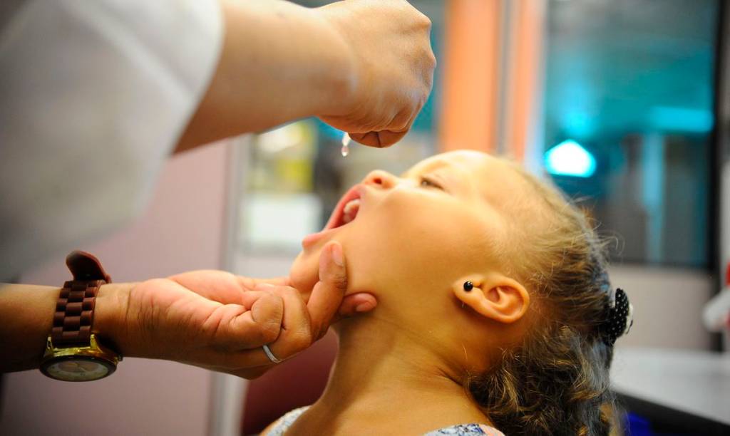 Imagem mostra criança sendo vacinada contra a poliomielite, abrindo a boca