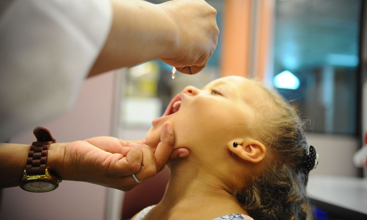 Imagem mostra criança sendo vacinada contra a poliomielite, abrindo a boca