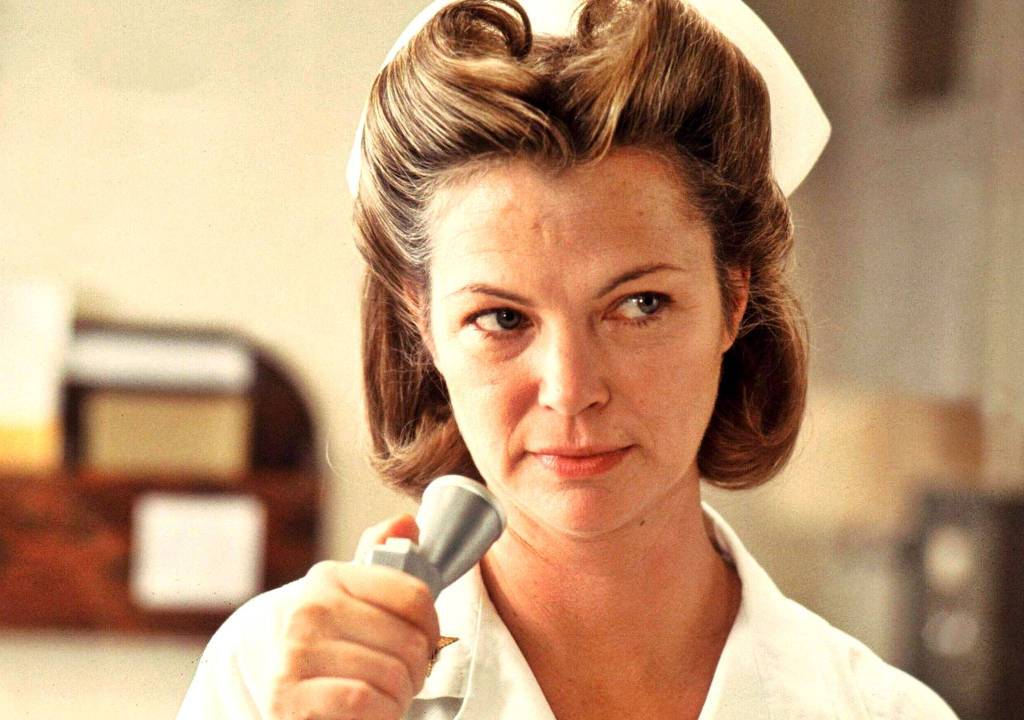 Louise Fletcher vestida de enfermeira segurando um microfone prateado.