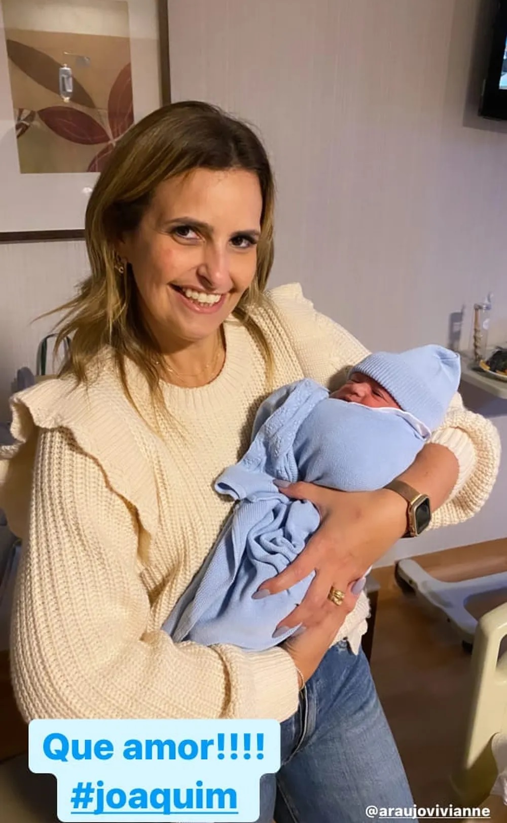 Moça loira sorri para a câmera e segura bebê envolto por roupinha de tricô azul-bebê.