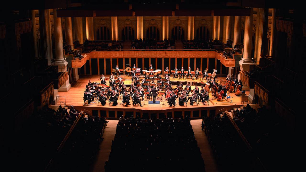 Imagem mostra sala de concertos com orquestra ao centro, no palco iluminado.