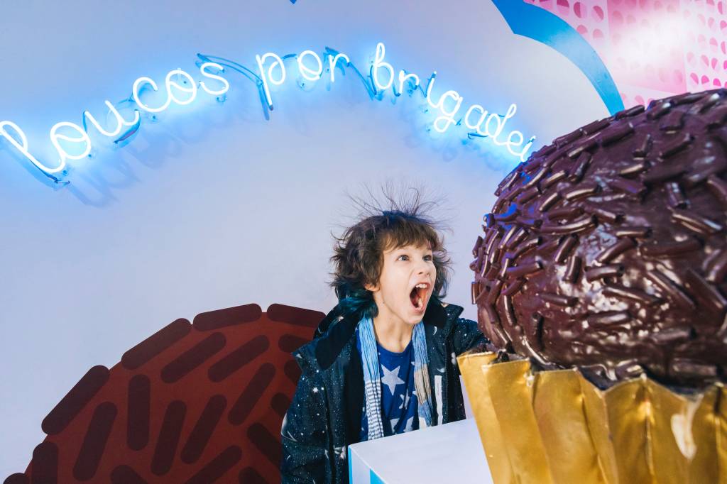 Criança grita em frente a brigadeiro gigante. Ao fundo, neon azul diz 