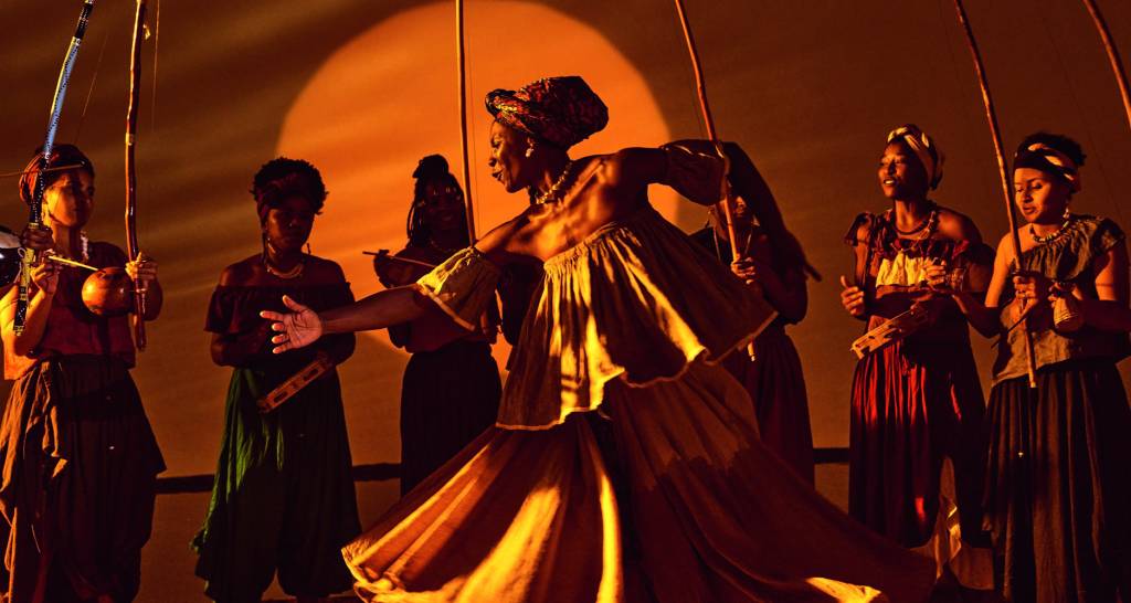 Imagem mostra mulheres dançando e tocando diversos instrumentos de percussão, iluminadas sob luz laranja