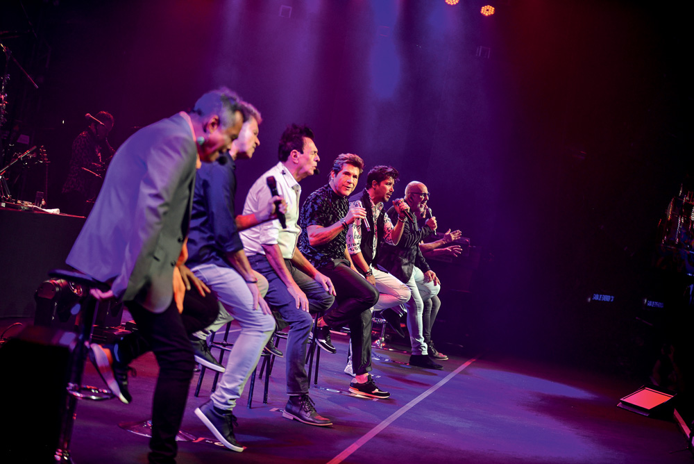 Imagem mostra diversos homens sentados em cadeiras, com microfones na mão, em cima de palco iluminado por luzes roxas