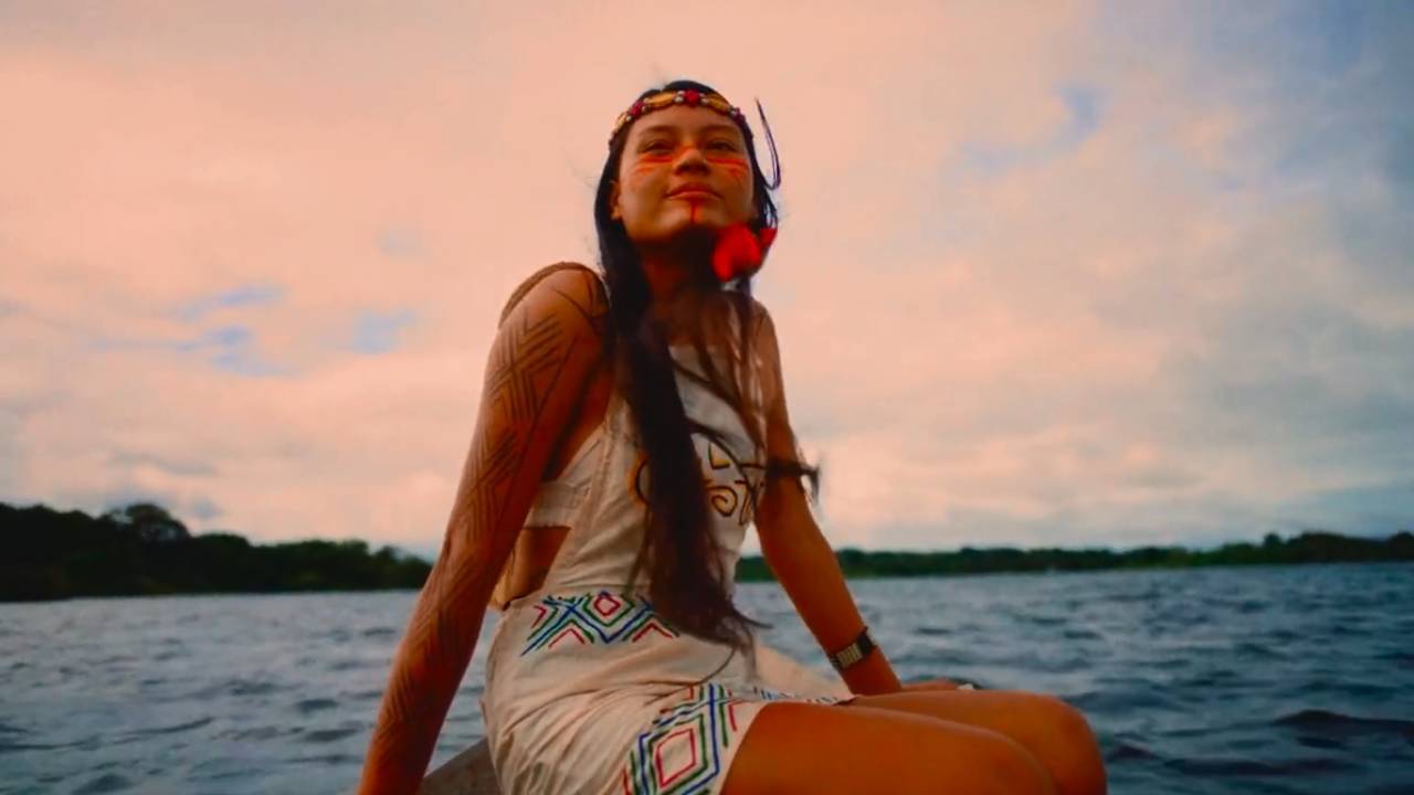 Moça indígena em barco aparece olhando para o horizonte com vestido branco. Ao fundo, nuvens alaranjadas e o lago.