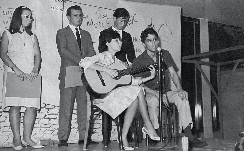 Entre os ex-alunos do colégio Santa Cruz, que fazem uma apresentação musical, está Chico Buarque cantando ao lado de uma moça que toca violão