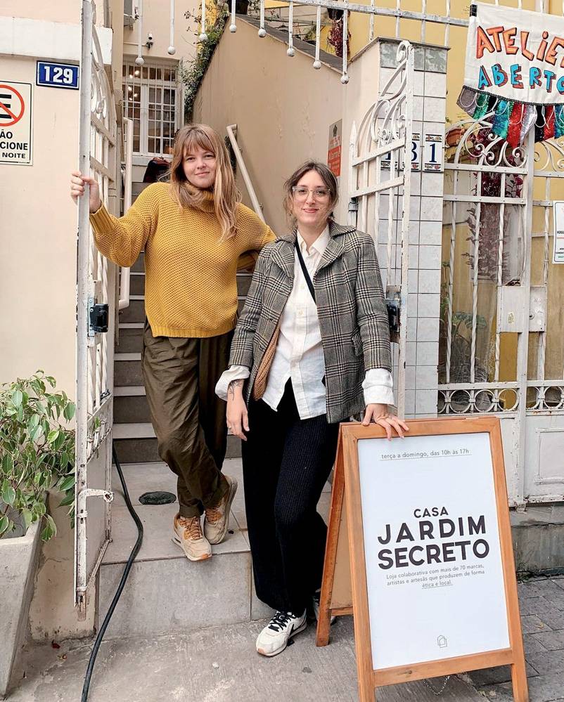 Claudia e Gladys, Jardim Secreto (Pamela Alves-Divulgação) Claudia e Gladys posam de pé em frente a escada que dá entrada a loja. Aparece no canto inferior direito uma placa no chão, onde Gladys apoia a mão, enquanto Claudia se segura no portão branco. Gladys usa óculos de grau, camisa branca, calça preta e blazer cinza. Claudia veste suéter cor mostarda e calça amarronzada.