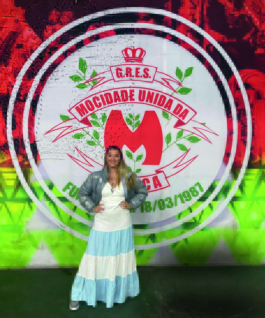 Crédito Arquivo pessoal_Divulgação.jpeg Imagem mostra mulher com vestido branco e azul, sorrindo em frente a cartaz de escola de samba