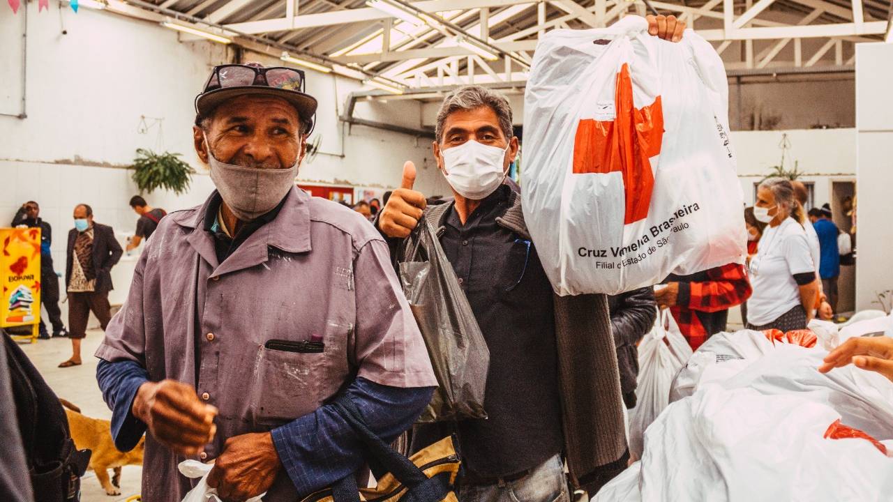 Dois homens de máscara seguram sacolas com cruzes vermelhas em galpão de teto branco.