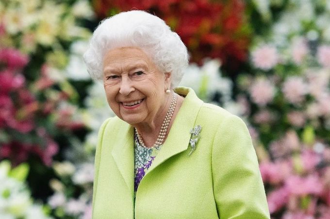 Rainha Elizabeth posa sorrindo para a câmera, usando terno verde claro, luvas e uma bolsa. Ao fundo, flores desfocadas