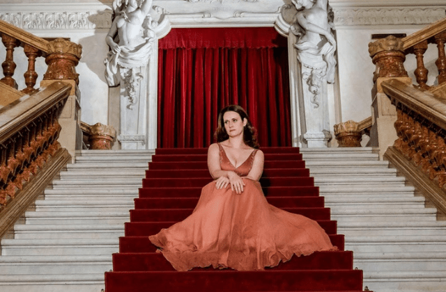 Imagem mostra mulher de vestido sentada em escadaria de mármore, com tapete vermelho