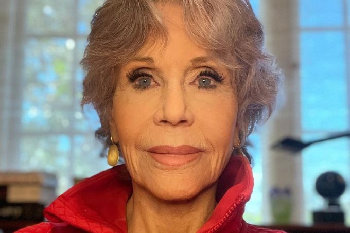 Jane Fonda posa com semblante neutro, usando blusa vermelha de gola alta. Ao fundo, vidro azul