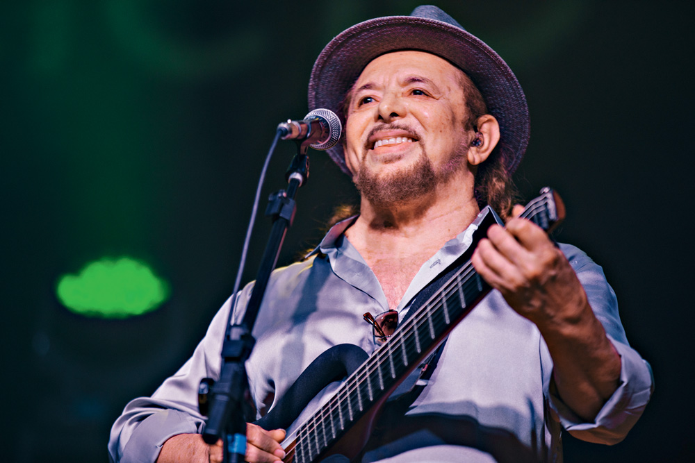 Imagem mostra homem de chapéu, segurando violão, em frente a microfone, cantando