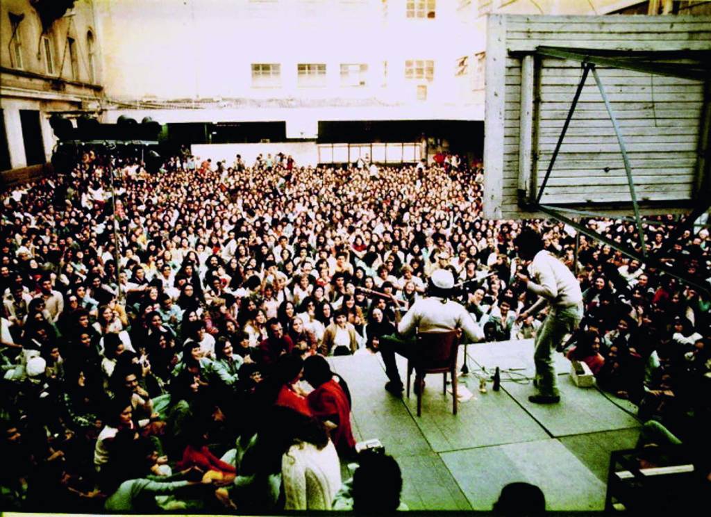 Imagem mostra pátio de escola lotado ao redor de pequeno palco com homem, tocando violão, sentado