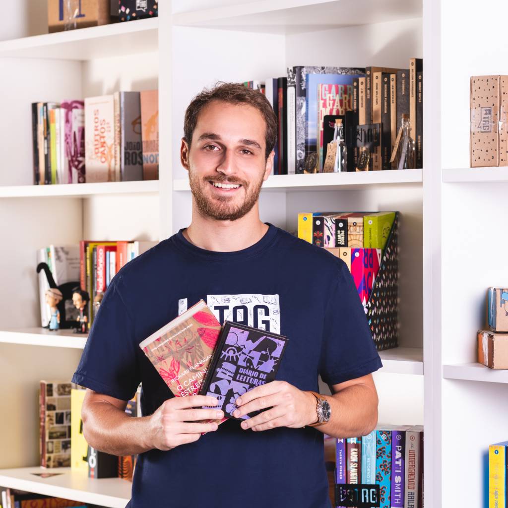 Gustavo Lembert sorri com dois livros na mão e uma camiseta azul-marinho com o logotipo TAG. Ao fundo, estantes brancas repletas de livros.