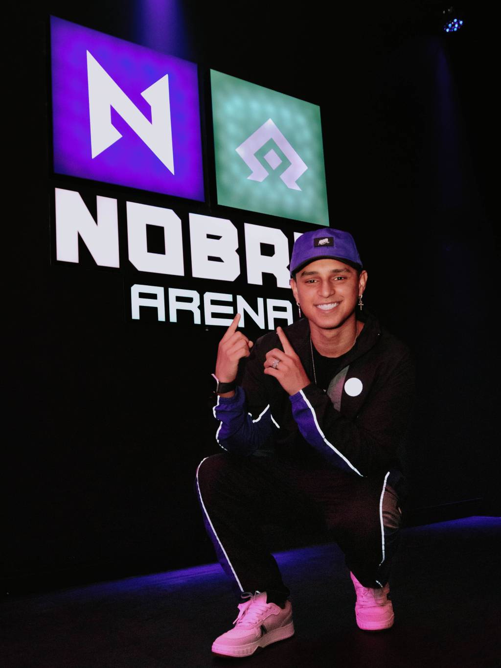 Gamer Nobru posa agachado, sorrindo e apontando para placa que está às suas costas, escrito Nobru Arena