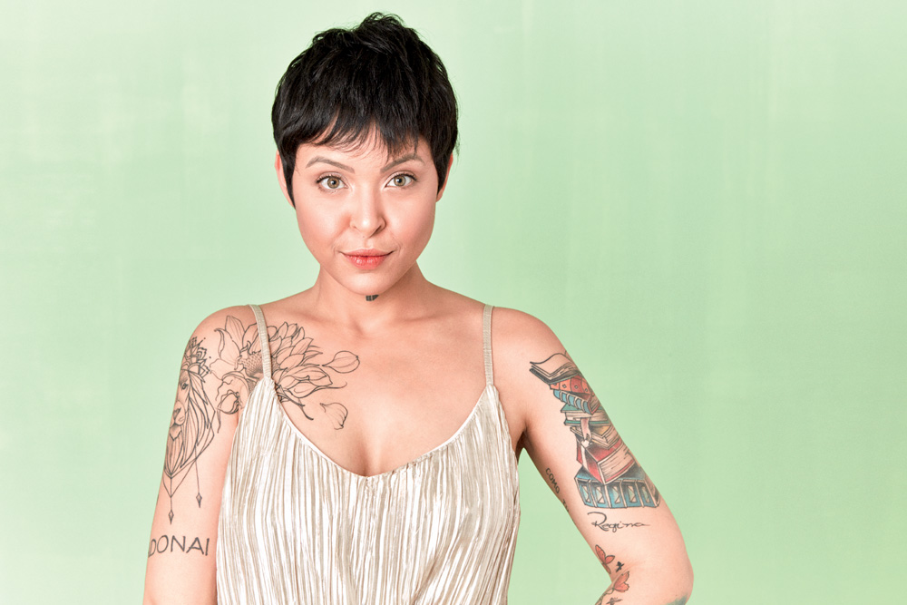 Imagem mostra mulher de cabelo preto, curto, com os braços tatuados, sobre fundo verde claro
