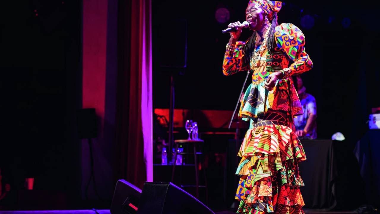 Imagem mostra mulher cantando em palco, com vestido longo e colorido
