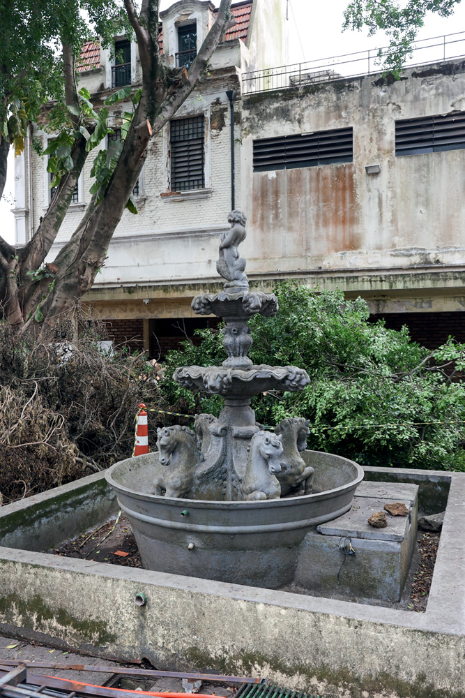 RESTAUROCOLEGIO50.jpg Imagem mostra fonte em área externa, perto de árvore. Ela é cinza e tem formatos de cavalos e anjos.