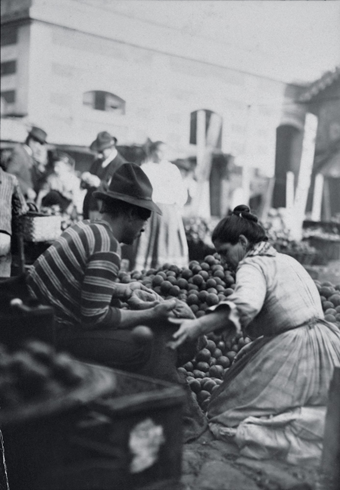 Imagem em preto e branco mostra mulher e homem pegando frutas em feira ao ar livre