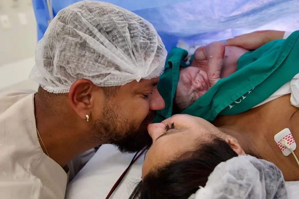 Viviane e marido se beijam com bebê recém-nascido nos braços.