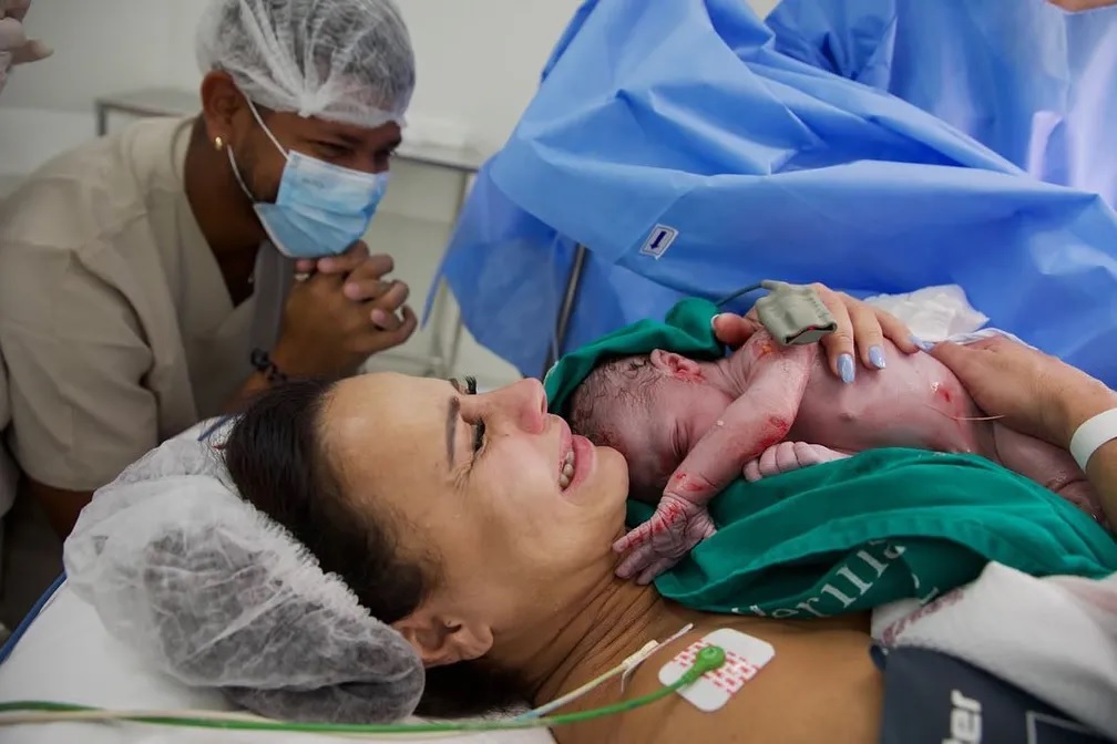 Viviane Araújo chora deitada com bebê recém-nascido nos braços. O pai aparece de máscara e touca ao fundo.