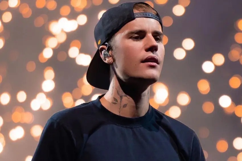 Imagem mostra Justin Bieber de boné e camiseta preta, olhando para o lado direito, com luzes ao fundo.