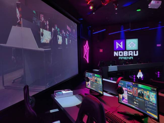 Imagem tirada da lateral do palco mostra computadores ligados, no primeiro plano, e ao fundo telão de LED e placa escrita Nobru Arena