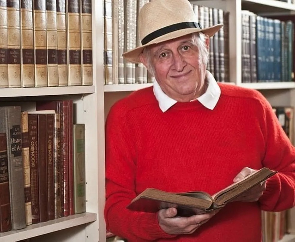 Sílvio Lancellotti posa sorrindo, usando chapéu e suéter vermelho, segurando livro aberto, diante de estante de livros.