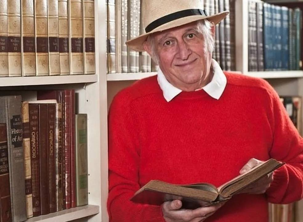 Sílvio Lancellotti posa sorrindo, usando chapéu e suéter vermelho, segurando livro aberto, diante de estante de livros.