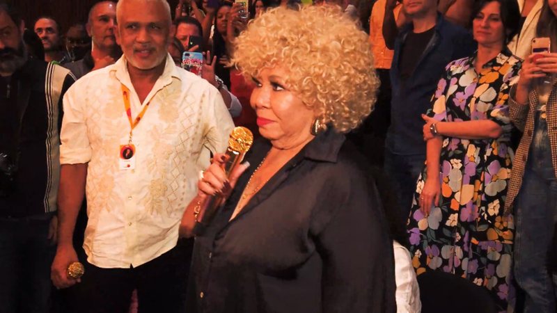Alcione, uma senhora idosa negra e com cabelos cacheados e grisalhos, segura um microfone dourado na plateia do teatro