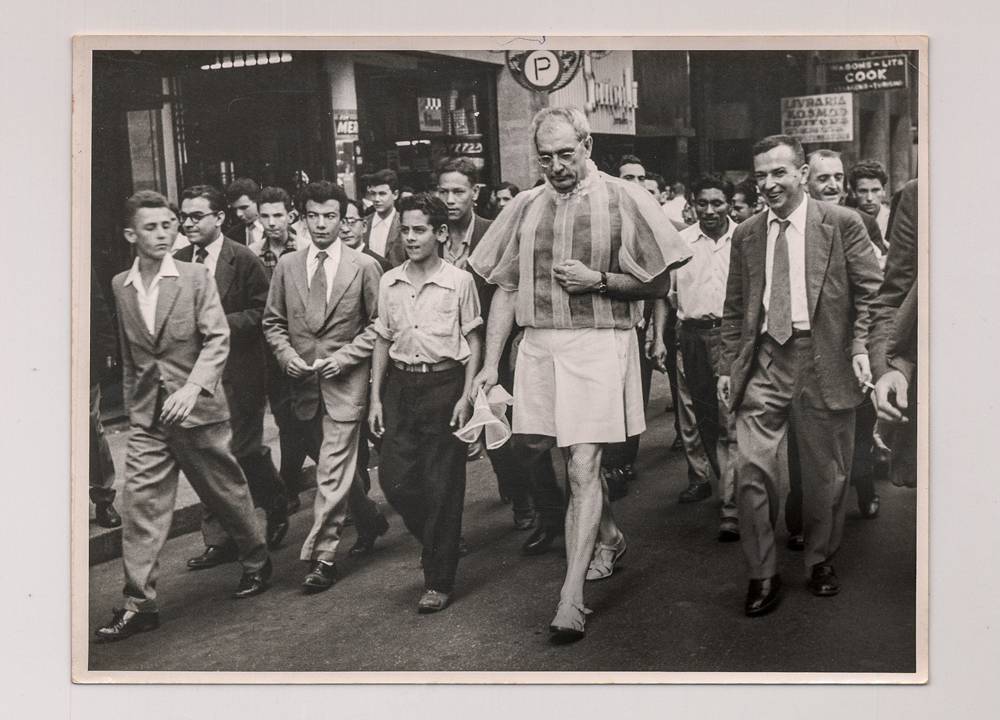 Imagem mostra foto em branco e preto de homem, de saia, andando em rua, com uma multidão ao seu redor