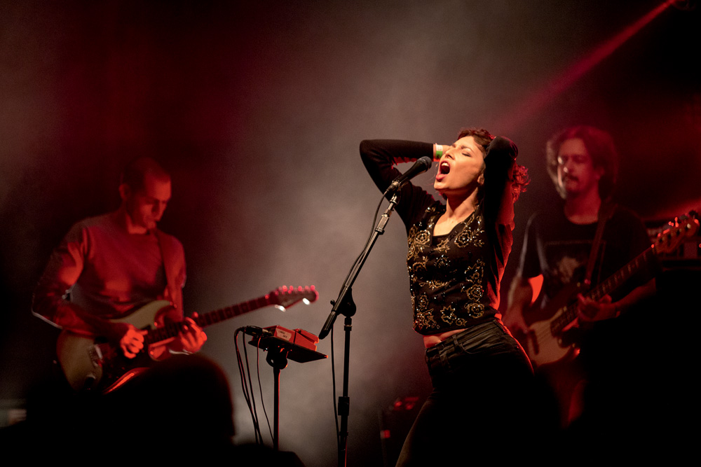 Imagem mostra palco com vocalista e outros dois músicos ao redor. A cantora está com a boca aberta e as mãos sobre a nuca