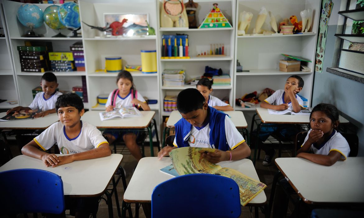 Foto mostra cinco alunos sentados em carteiras dentro da sala de aula.