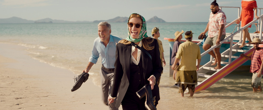 Imagem mostra mulher de terno e bandana, segurando salto, caminhando em praia e sorrindo