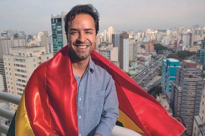 Ricardo Sales aparece sorrindo, com uma bandeira LGBT junto ao corpo e a cidade de São Paulo ao fundo