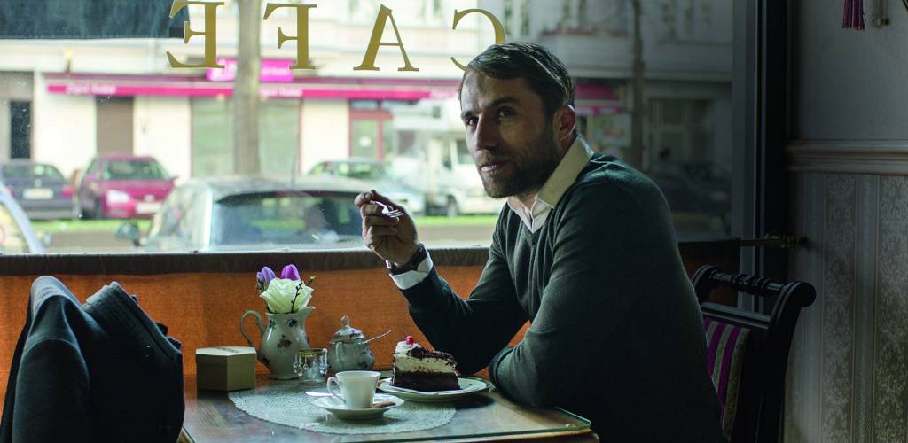 sp food festival – credito Karma Films.jpg Imagem mostra homem de suéter sobre camisa branca, tomando um café e comendo um bolo