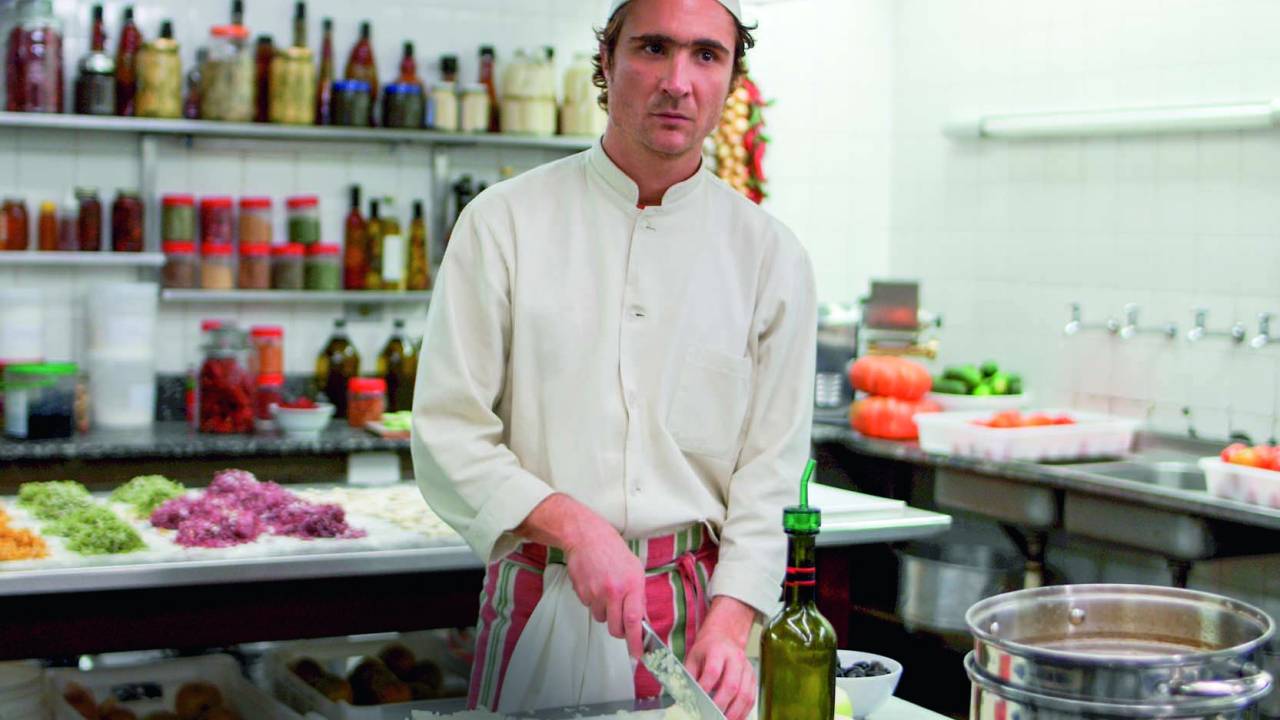 Imagem mostra cozinheiro, vestido de roupa branca e chapéu, picando cebola em cozinha