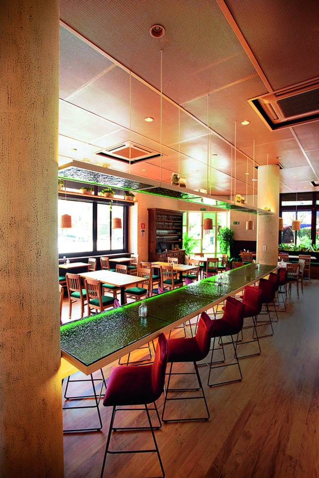 Ambiente interno de restaurante com mesas de tampo verde, amplas janelas e pilastras, sem nenhuma pessoa sentada às mesas