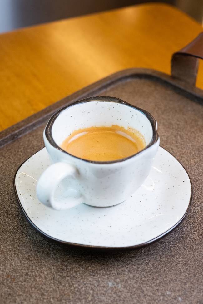 xícara branca com borda preta contendo café expresso sobre pires branco em uma bandeja marrom apoiada em uma superfície de madeira