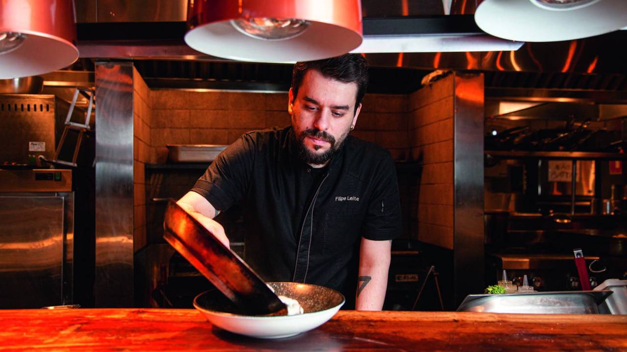 Chef Filipe Leite despeja conteúdo de uma panela em um recipiente sobre mesa de madeira em uma cozinha