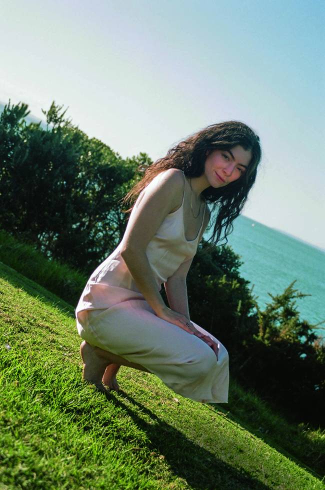 Foto de Lorde agachada em gramado, com mar ao fundo