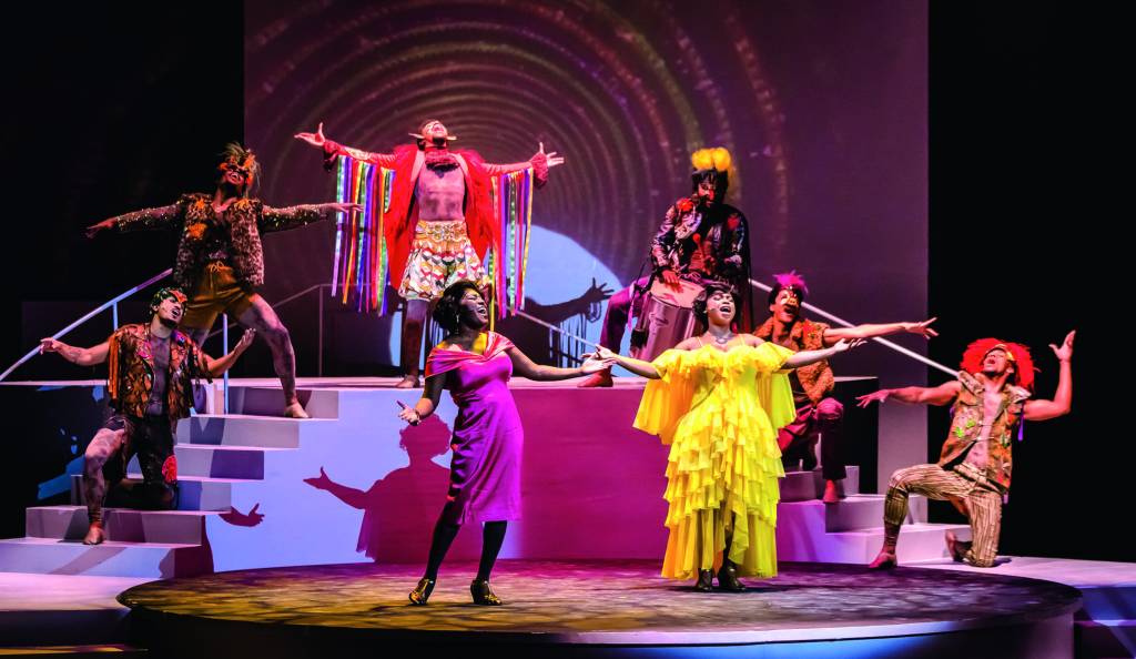 No palco, duas cantoras negras interpretam Alcione enquanto cantam. Uma usa um vestido amarelo bufante e a outra, um vestido midi rosa. Outros atores estão atrás, com figurinos coloridos