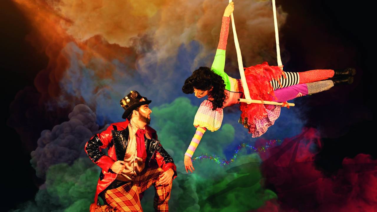 Um casal de atores veste figurinos coloridos de circo, em um fundo com nuvens coloridas. Ela se pendura em um trapézio e estende um dos braços para ele, que está agachado