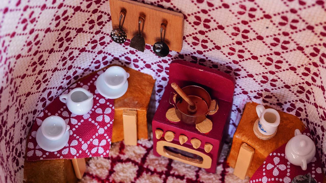 Miniatura de cozinha vista por centro. As paredes são estofadas com tecido estampado branco e vermelho. Há fogão, mesa e armário, com panelinhas e demais utensílios