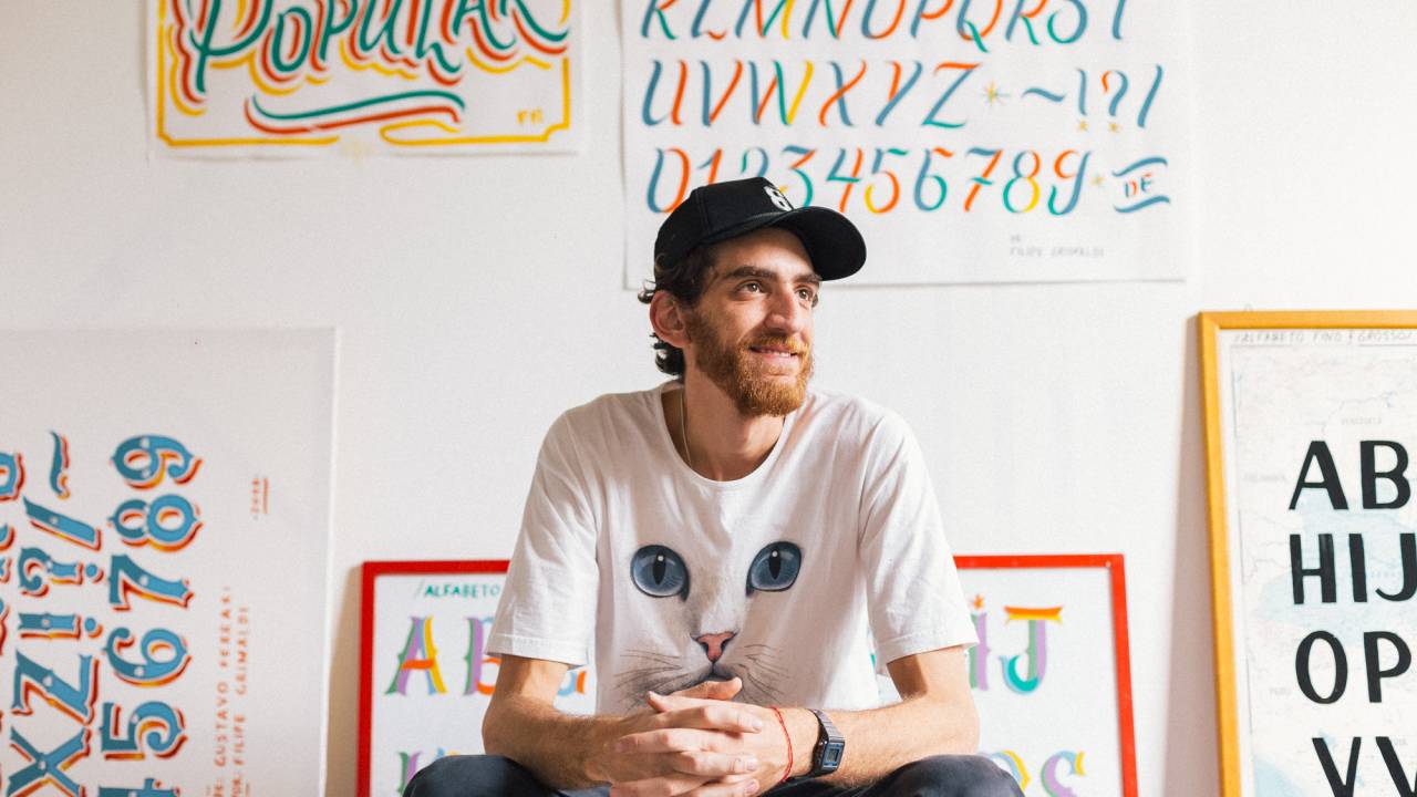 Filipe Grimaldi está sentado e sorri olhando para a direita. Veste camiseta branca, calça jeans e boné preto. Tem barba e ao fundo aparecem letras estilizadas na parede, uma delas formando a palavra 'popular'.