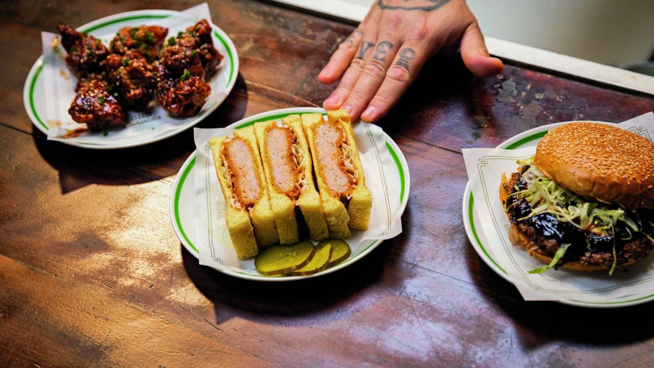 Sando, frango aka aague e sanduíche com molho teriyaki em pratos de sobremesa sobre guardanapos de papel, sobre mesa de madeira, com uma mão branca tatuada em posição de empurrar o prato do sando