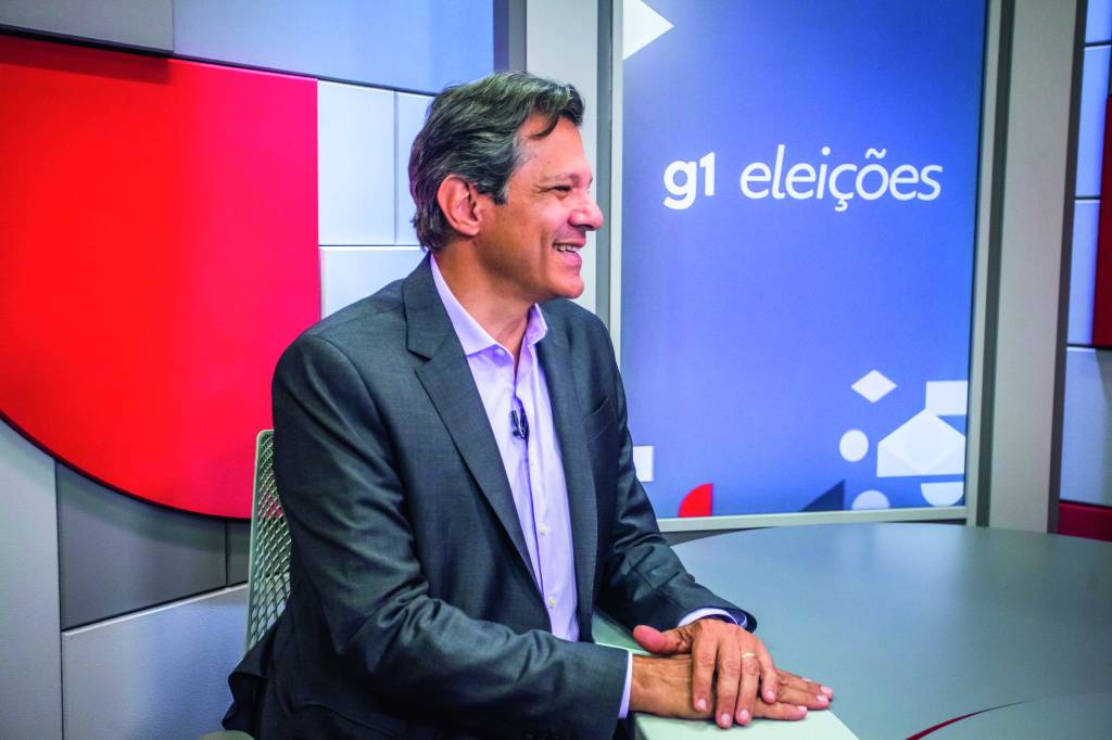 Fernando Haddad em púlpito durante debate eleitoral. Ele veste um terno e sorri. É fotografado de perfil