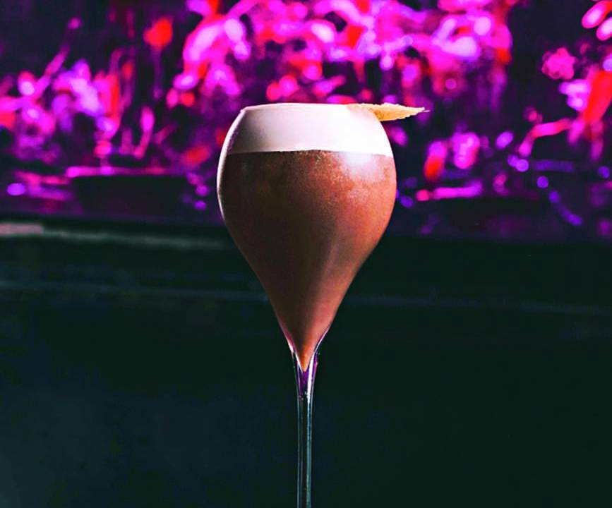 Taça alta contendo um drink arroxeado e opaco contra fundo escuro com luzes cor de rosa
